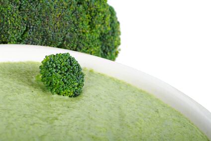 Een kom broccolisoep, geïnspireerd op een recept van Piet Huysentruyt met kaas in de soep verwerkt, met broccoli op de achtergrond